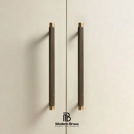 Two long cylindrical cabinet handles with a dark textured wrap and brass end caps, mounted on a light beige door. The Modern Brass logo is visible at the bottom.