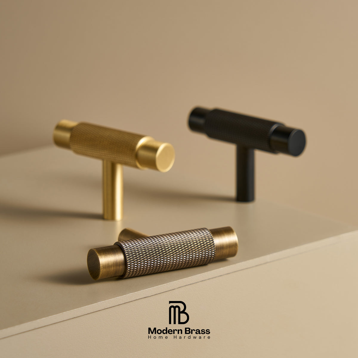 Three modern cabinet knobs by Modern Brass are displayed on a beige surface against a matching beige background. The set includes two T-shaped knobs in polished brass and one horizontal knurled mesh cylinder in a dark gunmetal/black finish.
