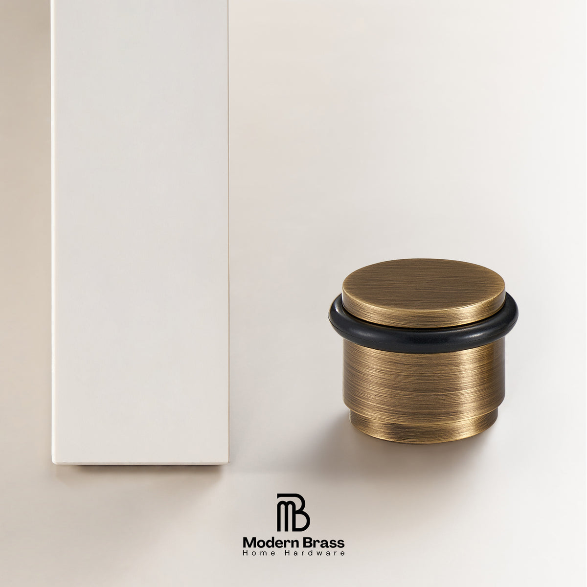 A close-up studio shot of a brass cabinet pull handle with a brushed antique brass finish and a black rubber gasket, shown next to its white rectangular packaging box on a clean off-white background. The Modern Brass logo is visible at the bottom.