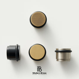 Three round cabinet knobs in black, matte brass, and brushed brass are arranged on a light beige surface against a warm beige background. The Modern Brass logo is visible at the bottom.