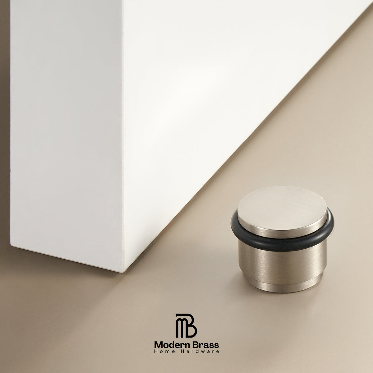 A brushed brass door stopper with a black rubber seal, shown on a beige floor next to a white cabinet corner, with the Modern Brass logo at the bottom.