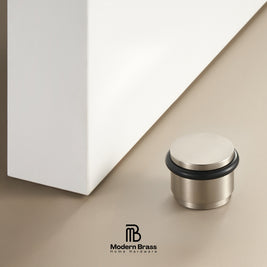 A brushed brass door stopper with a black rubber seal, shown on a beige floor next to a white cabinet corner, with the Modern Brass logo at the bottom.