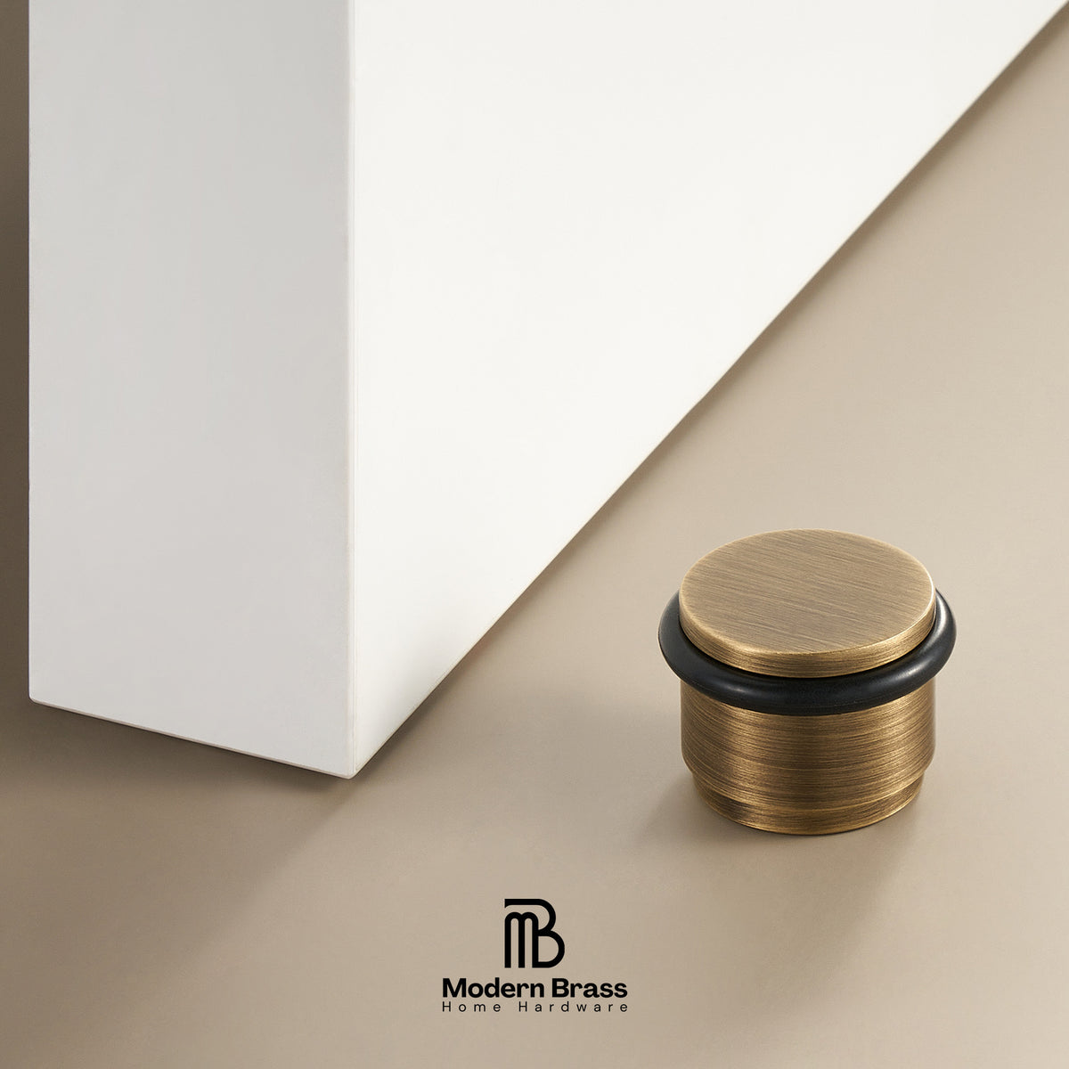 A close-up of a brass door stopper with a brushed gold finish and a black rubber ring, positioned next to a white furniture leg on a beige background. The Modern Brass logo is visible at the bottom.