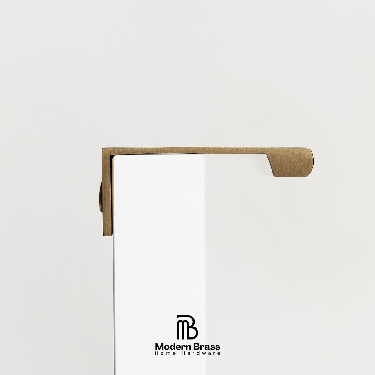 A close-up of a brass door handle mounted on a white door, showing the brushed metal lever and its L-shaped bracket. The Modern Brass logo is visible on the door.