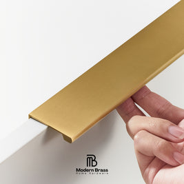 A close-up of a hand holding a brushed brass cabinet trim piece against a white wall, with the Modern Brass logo at the bottom.