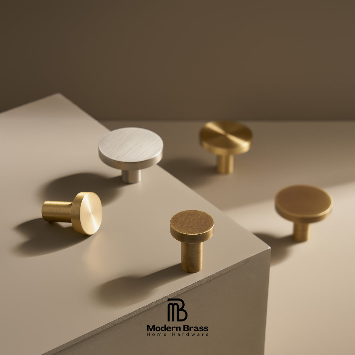 Four round cabinet knobs in brushed brass and matte white finishes displayed on a beige geometric pedestal, with the Modern Brass Home Hardware logo at the bottom.