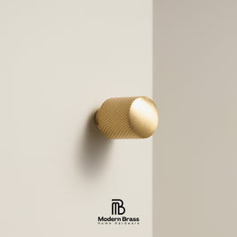 A modern cylindrical brass cabinet knob with a textured knurled surface and a smooth polished end, mounted on a minimalist beige wall. The Modern Brass logo is visible at the bottom.