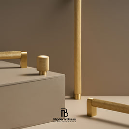 Modern brass home hardware set including textured cylindrical knobs and bar pulls in a warm brushed gold finish, displayed on a minimalist beige background with a geometric pedestal.